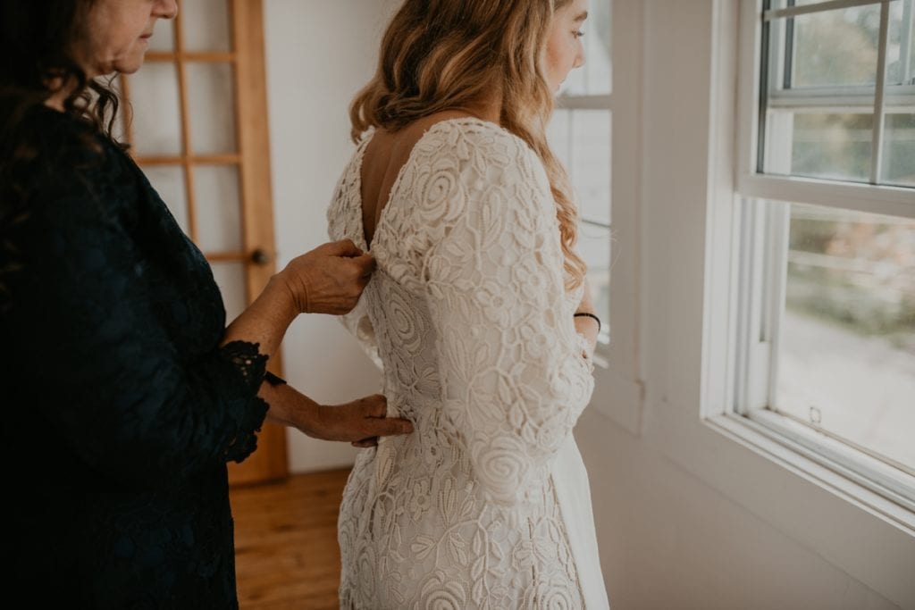 Getting ready bridal portrait. Bride's mother is zipping up her lace dress as the bride is looking out a window. Captured at The Clearing Wedding venue in Shedden, ON