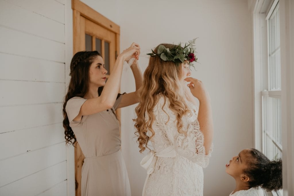 Getting ready photo of the bride at The Clearing in Shedden, ON. A bridesmaid is tying the back of her boho-inspired floral crown. The flower girl is looking up at the bride.