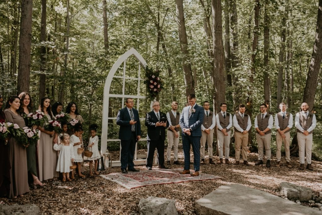 The Groom is standing at the wedding altar in a forested area at The Clearing in Shedden, ON. He is looking down at his hands waiting for the bride. The wedding party is lined up beside him.