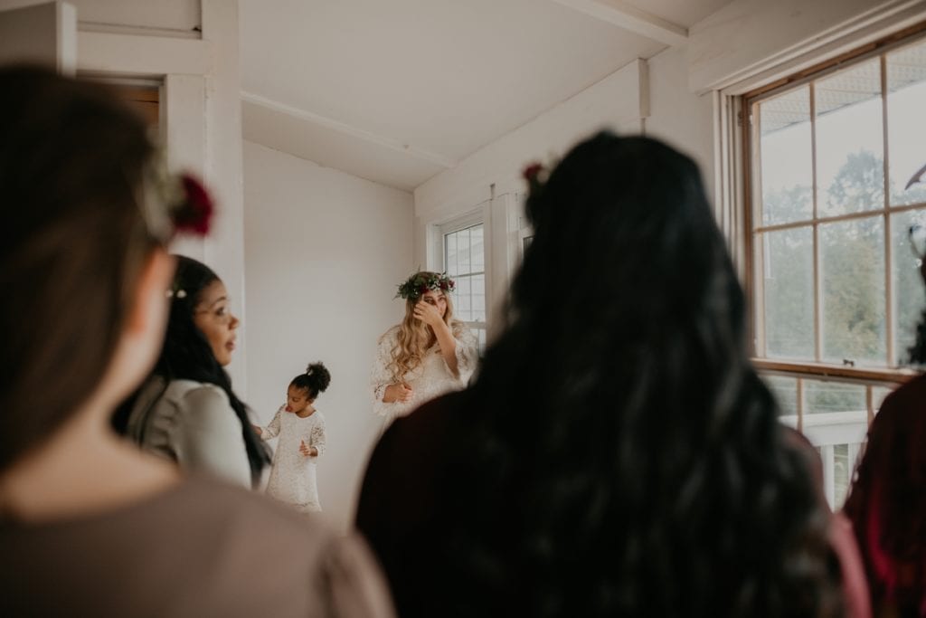 The bride is facing her bridal party as they see her dressed for the first time. In the foreground of the image is the bridesmaids backs and the bride is standing between their heads wiping a tear from her eye. Wedding venue is The Clearing in Shedden, ON.