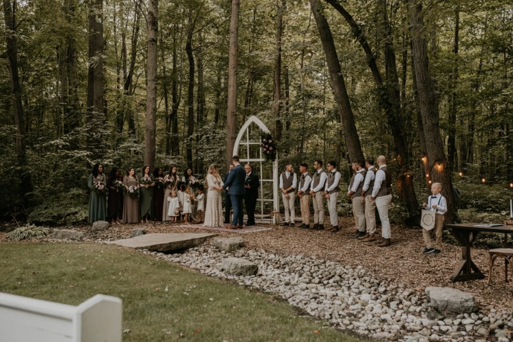 Bride and groom hold hands at their wedding altar at The Clearing in Shedden, ON. Their bridal party stands on either side of the bride ad groom.