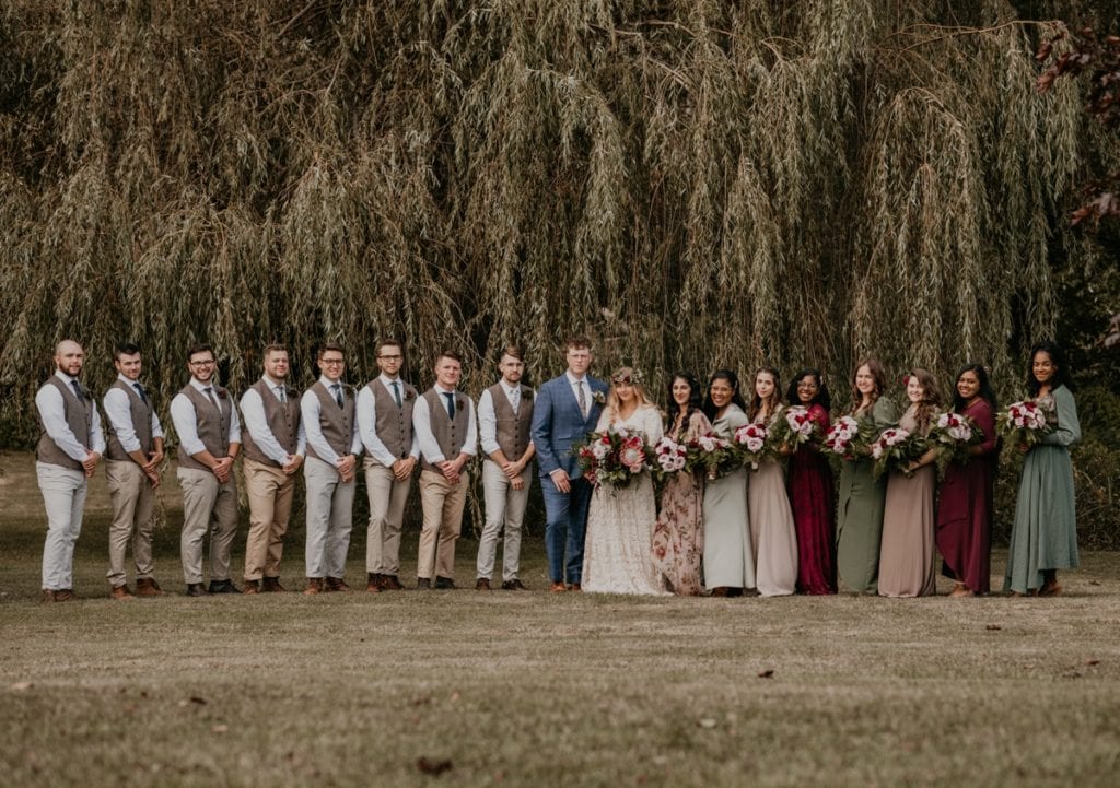 Posed wedding party portrait. The bride, groom, and wedding party are lined up underneath a willow tree at The Clearing in Shedden, ON