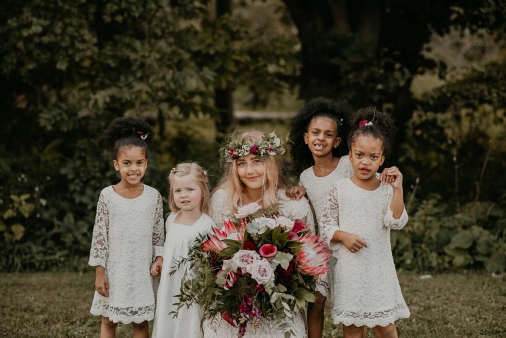 The bride is crouching down for a photo with her flower girls at The Clearing in Shedden, ON. The bride is holding a large flower bouquet.