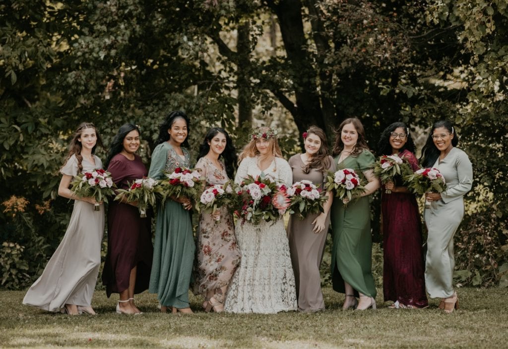 The bride is posing with her bridesmaids for a wedding day portrait at The Clearing in Shedden, ON. The bridesmaids are in assorted beige, green, and burgundy hues, all holding large floral bouquets. The are smiling at the camera and the wind is gently blowing their hair and dresses. Captured by top London, Ontario wedding photographer Ashlee Ellison.