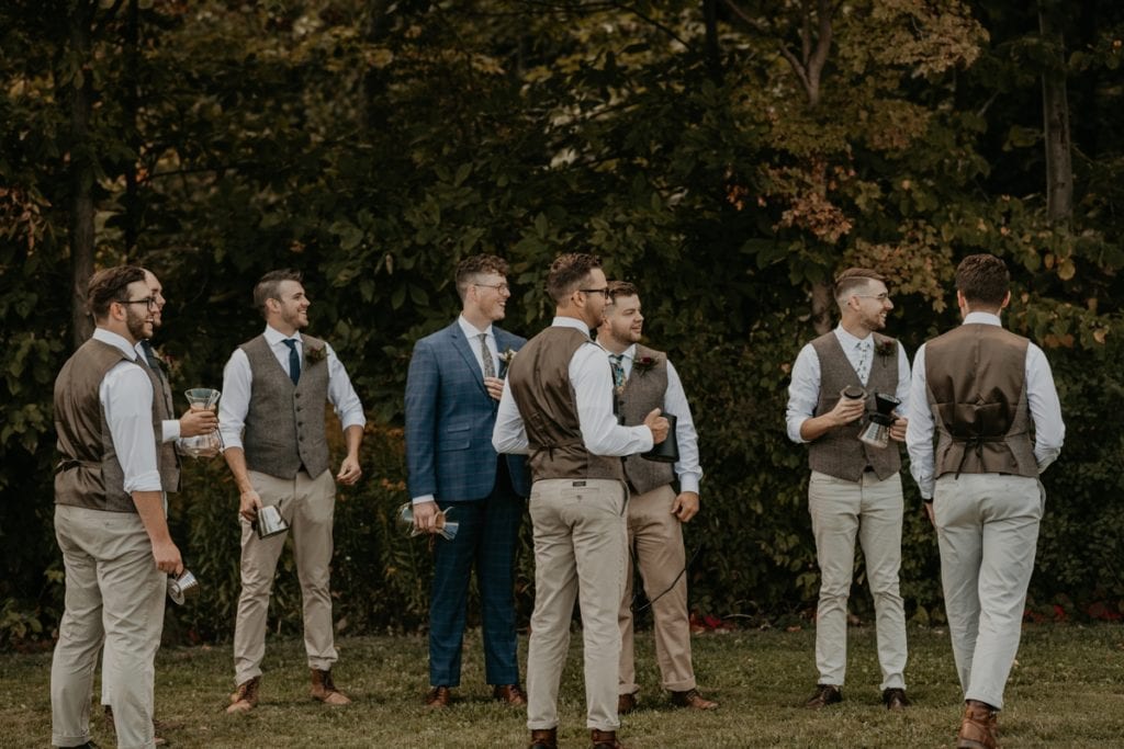 The groom and his groomsmen are standing outdoors having a conversation and laughing at The Clearing in Shedden, ON.