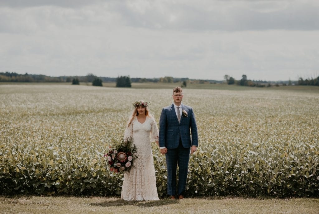 Bride and groom stand hand-in-hand in front of a farmer's field for a wedding day photo. The bride is holding a large oversize bouquet.
