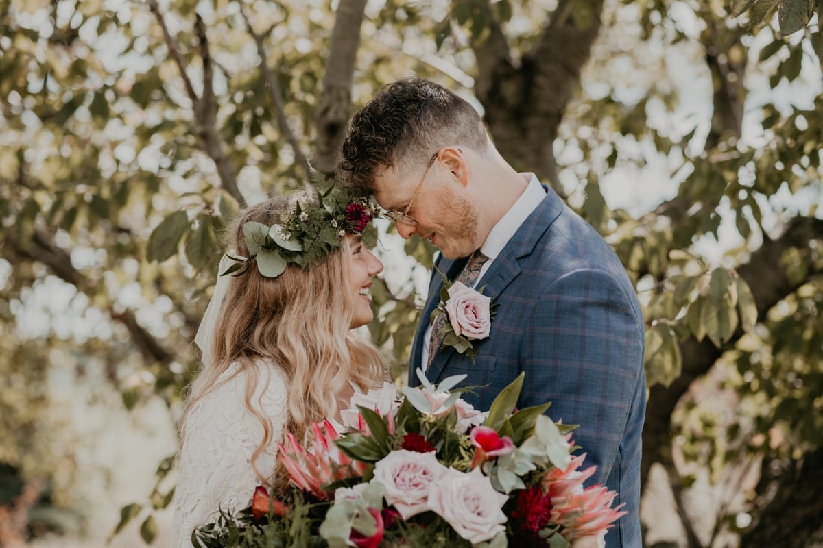 Bride and groom are facing each other and smiling. Their heads are touching for an intimate wedding day portrait. Bride is wearing a floral crown and holding an oversized bouquet in front of her. Captured by best London, Ontario wedding photographer Ashlee Ellison.