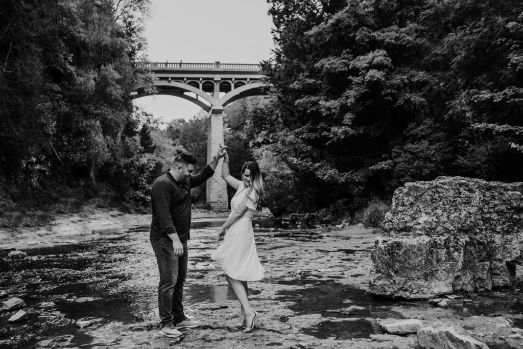 Man and woman are standing on a large rock in the middle of a low-lying rocky stream. The man is twirling the woman and the woman is playing with her dress. the man is looking at the woman and the woman is looking over her shoulder. behind them is a very tall stone bridge and lush greenery on either side of the stream. Captured by top london ontario wedding and engagement photographer ashlee ellison.
