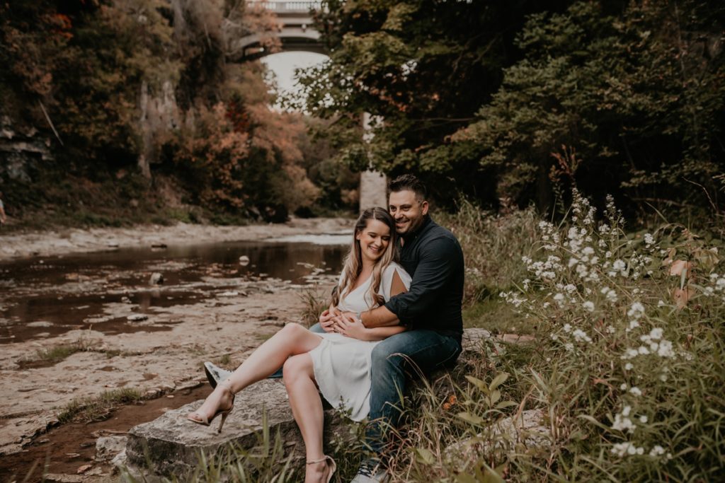 Couple pose along side a river for a candid engagement photo. They are sitting on a rock. the woman is sitting with her back to the man's chest. The man's arms are wrapped around her and her hands are atop of his. Behind them is a very tall stone bridge and lush fall seasonal greenery. Captured by best london ontario wedding and engagement photographer ashlee ellison.