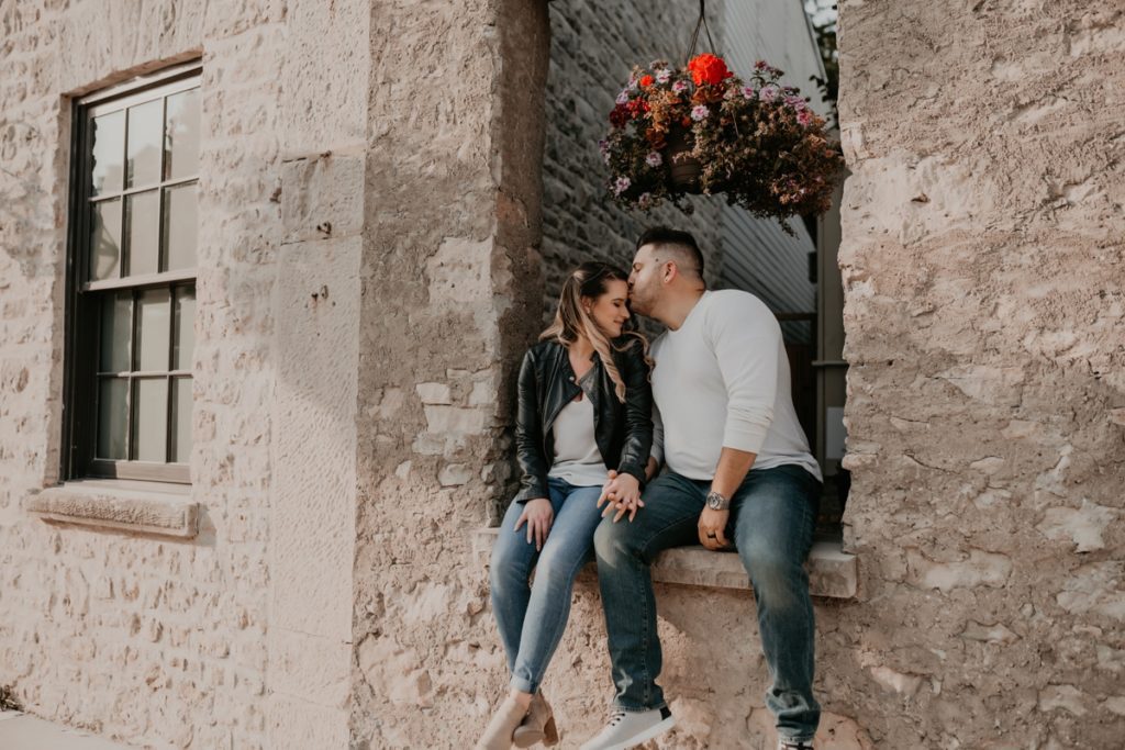 Couple sit on a rustic window ledge holding hands. the man is kissing the woman's forehead. both of them have their eyes closed. Above them is a planter with bright red and purple hues. Captured by top london ontario engagement photographer ashlee ellison.