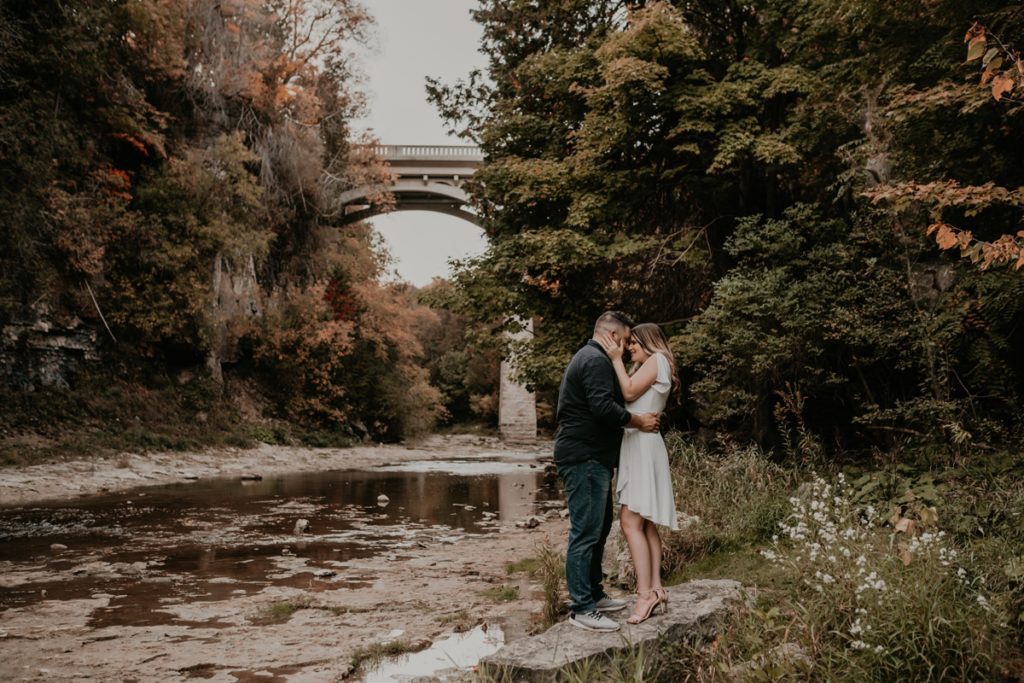 Couple pose for their London, Ontario Engagement session. They are standing on a large rock beside a low-laying stream in a gorge. Behind them is a very tall stone bridge. Both sides of the river are surrounded by lush greenery beginning to turn colours for the fall season. Captured by top London Ontario wedding and engagement photographer ashlee ellison.