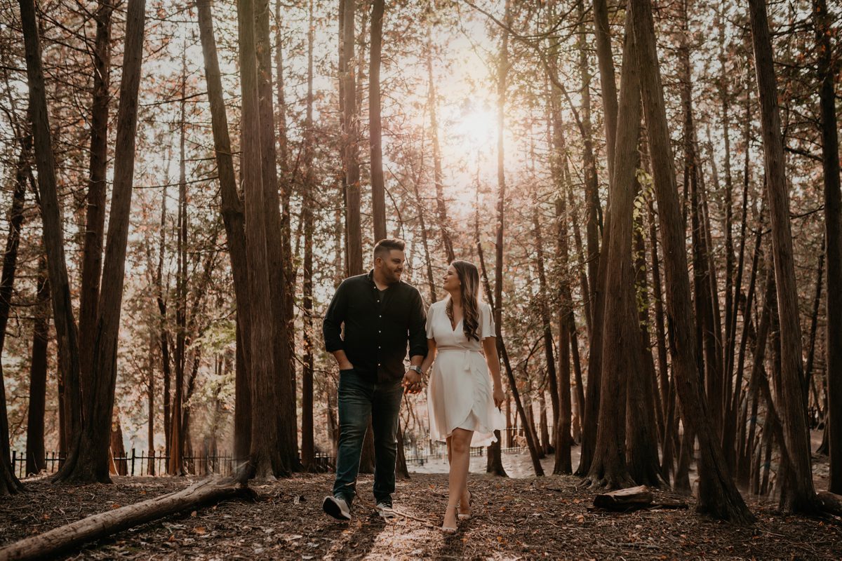 Couple walk through a forested area during golden hour. They are holding hands and smiling at each other. The sun is setting behind them creating a golden hue.