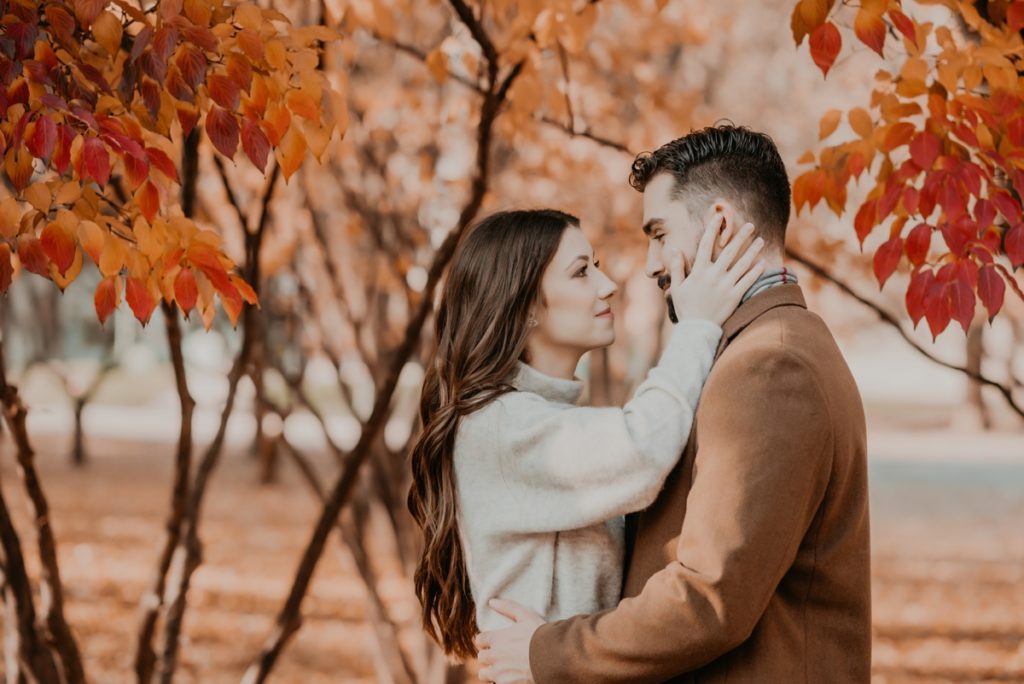 close up image. Man and woman stand amongst red fall foliage at UWO in London, ON for their engagement session. The man's arms are around the small of the woman's back. The woman's hands are touching his cheek. They are looking into each other's eyes. Captured by top london ontario wedding photographer ashlee ellison.