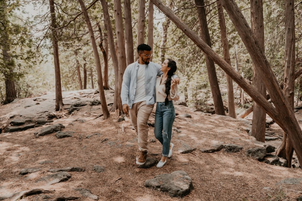 Man and woman walk through forested area for a candid engagement photo. Man's arm is around the woman's shoulders and she is reaching up to hold his hand. They are looking at each other and smiling. Captured by top london ontario engagement photographer ashlee ellison.