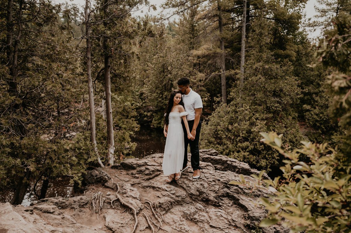Man and woman stand atop of a large rock with tree roots growing overtop of it surrounded by lush forest greenery. The woman's back is to the man's chest. They are holding hands. She is looking over her shoulder and he is gently kissing the top of her head. Captured by top london ontario engagement photographer ashlee ellison.