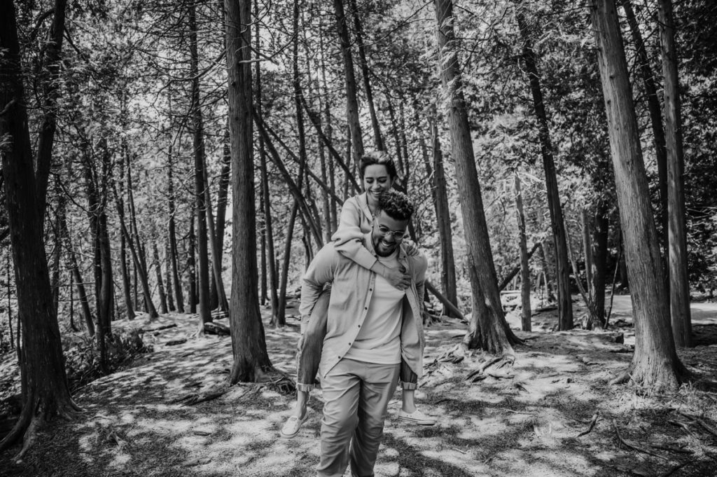 man piggybacks woman through forested area for a fun and candid engagement photo. The woman's arms are gently draped around the man's neck. They are both smiling and looking down toward the ground. captured by top engagement and wedding photographer in london ontario ashlee ellison.