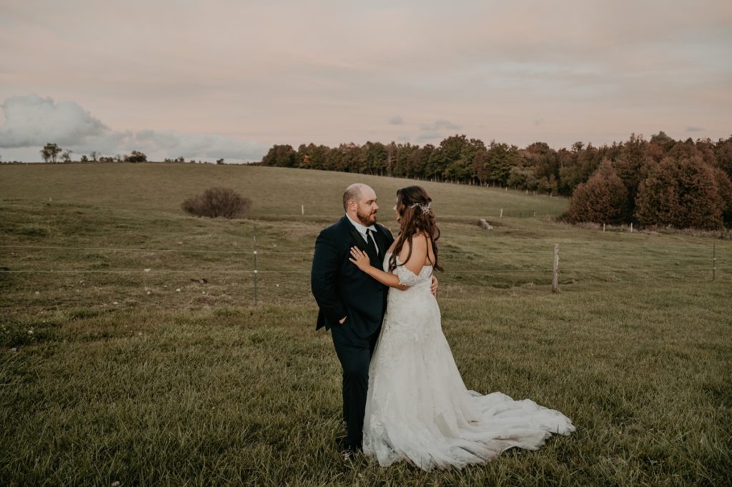 Bride and groom stand in a manicured grassy area at Luso Valley Estate Farm. Behind them are rolling fields of grass and evergreen trees. The bride and groom are standing chest-to-chest and looking at each other.