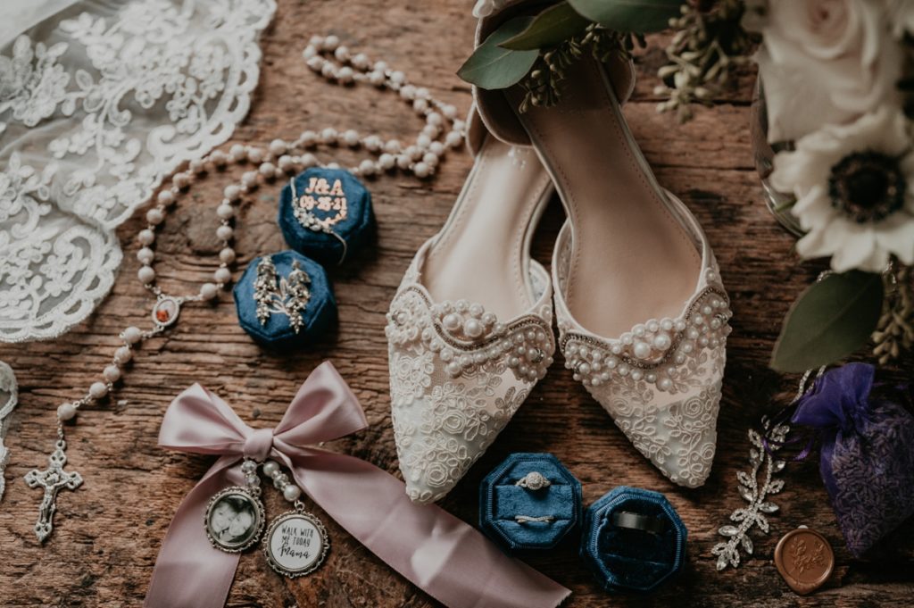 Flat lay details image featuring pearl and lace embellished shoes the brides jewelry, a rosary, and a pink bow to be affixed to her bouquet Attached to the bow are two tiny memory lockets, one with a picture of the bride and her deceased mother, the other with an inscription that says "walk with me today mama". Photographed atop rustic distressed wood at the Luso Valley Estate Farm in Orangeville, Ontario.