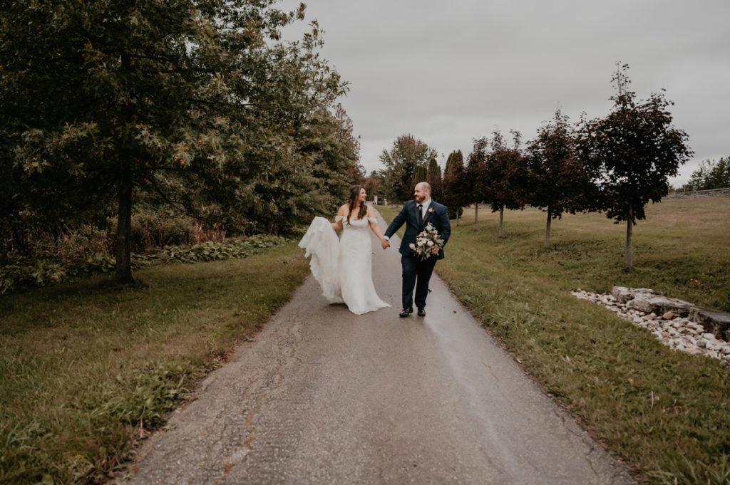 Bride and groom walk hand in hand along a paved pathway a the Luso Valley Estate Farm for their Orangeville ontario wedding photos. The bride is holding her gown and the groom is holding her flowers. They are looking at each other.