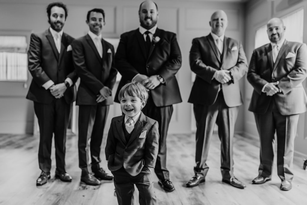 Black and white image. Ring bearer laughs with his hands in his pockets as the room and his groomsmen are behind him, blurred and smiling. Captured at Luso Valley Estate Farm wedding barn.