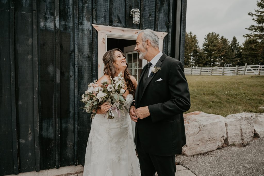 Bride and her father share a laugh before her Luso Valley Estate Farm wedding. They are standing in front of the barm, looking at each other and laughing.