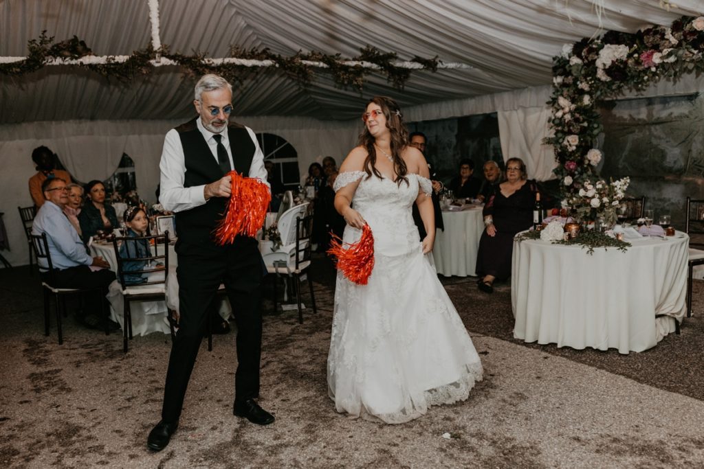 Bride and her father share a fun choreographed dance at her Luso Valley Estate Farm wedding reception. The bride and her father are both wearing tinted glasses and each holding a red pompom.
