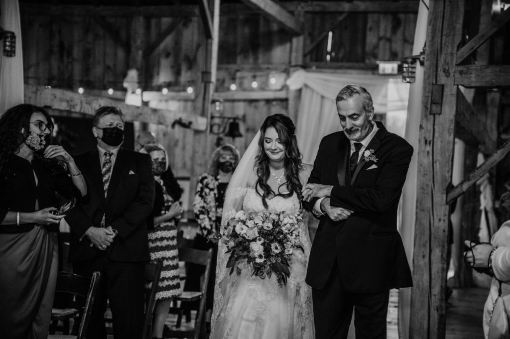 Brides father walks her down the aisle at the Luso Valley Estate Farm barn. Her arm is draped over her father's right arm and his left hand is atop of hers. Both are looking down, smiling deep in thought as the bride's mother has passed.