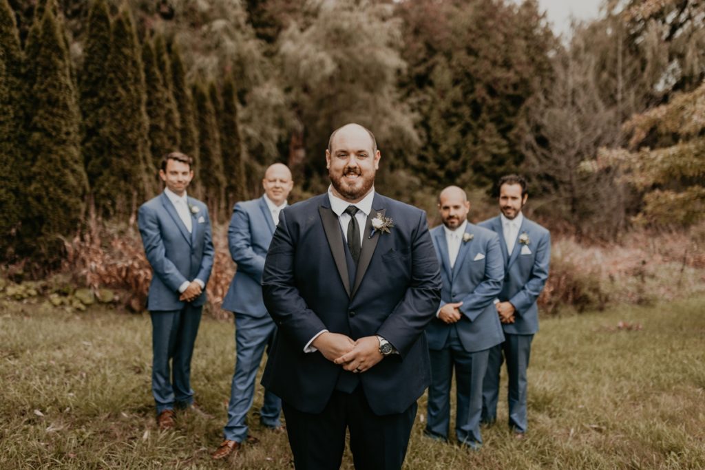 Groom stands smiling facing the camera with his hands folded in front of him as his groomsmen look on in the distance. Captured at Luso valley Estate Farm.