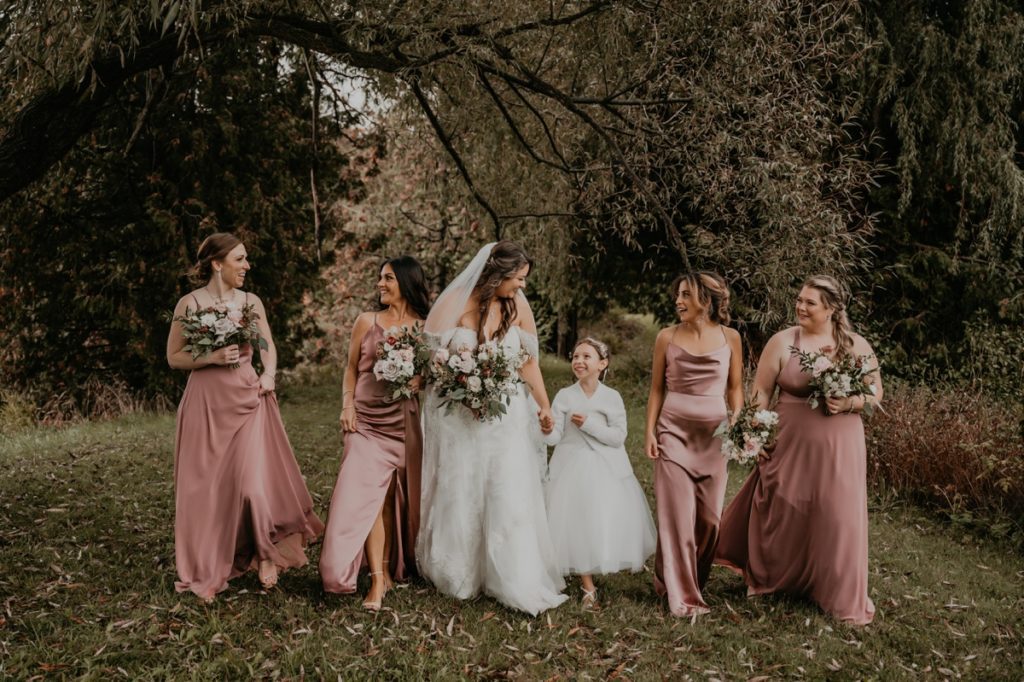 Bride, 4 bridesmaids, and flower girl walk on a grassy area of the Luso Valley Estate Farm for an informal wedding portrait. All are smiling and looking at each other, the flower girl holding the bride's hand. Behind them in a weeping willow tree.