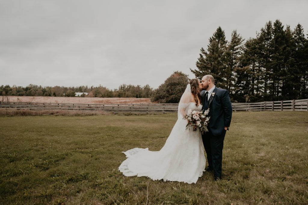 Bride and groom share a kiss on the grounds of the Luso Valley Estate farm in Orangeville, Ontario. Behind them off in the distance are farm fencing and wheat.