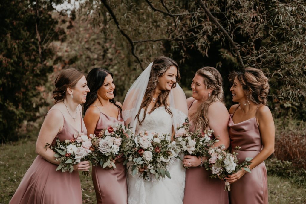 Bride and her four bridesmaids, wearing dusty pink, pose for an informal wedding day photo. They are standing in a line, bride in the middle, with their bouquets in front of them. They are all smiling and laughing at each other. Captured on the grounds of the Luso Valley Estate Farm in Orangeville by top Ontario wedding photographer Ashlee Ellison.