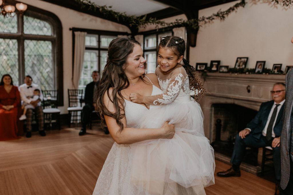 The bride and her daughter share a dance and her Elsie Perrin Williams Estate Wedding reception in London, Ontario as guests look on and smile. The bride is carrying her daughter, approx age 5. The bride is smiling at the little girl and the little girl is laughing.