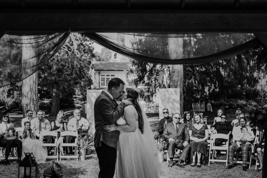 Bride and groom share their first kiss at their Elsie Perrin Williams Estate Wedding. The image is captured from behind the altar so you can see the bride and groom share their first kiss and the expressions on the wedding guests.