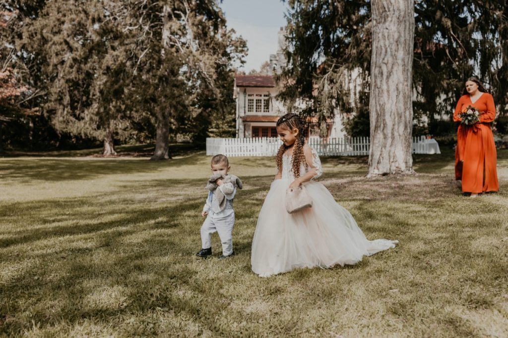 Flower girl and ring bearer walk along the grass on their way to the outdoor wedding ceremony on the grounds of the Elsie Perrin Williams Estate in London, Ontario. In the background, the building's white stucco facade and historic windows are featured.