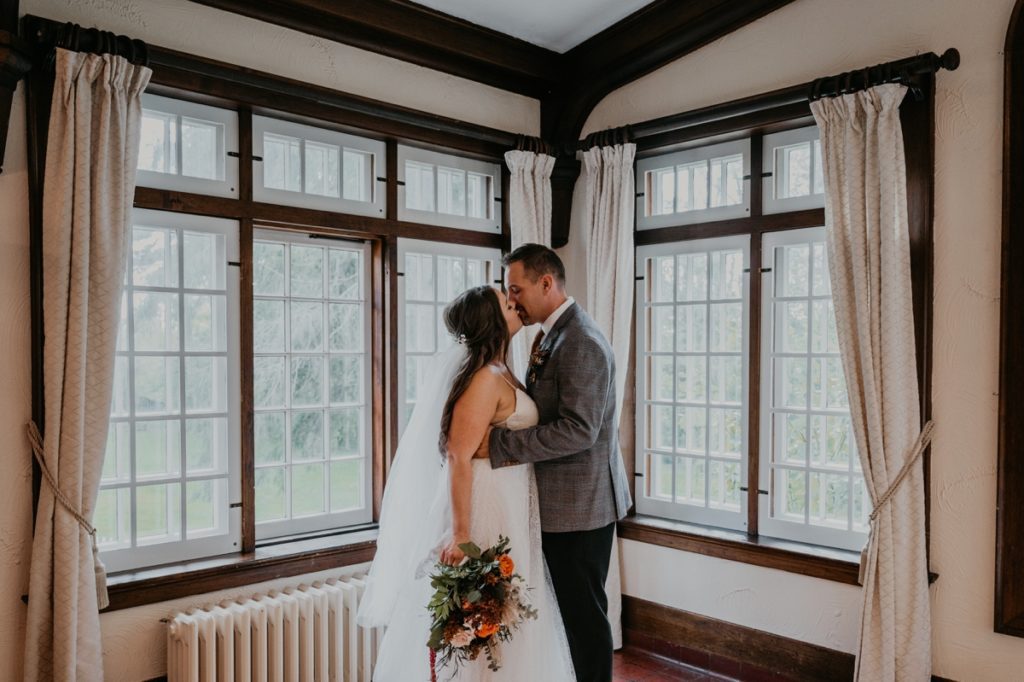 Bride and groom share an intimate kiss in front of the historic windows as the Elsie Perrin Williams Estate before their wedding reception.