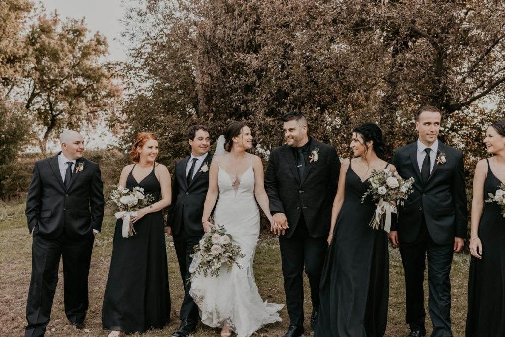 Bride and groom are holding hands and they walk through a grassy area for a candid wedding day photo. Bride and groom are smiling at each other and the wedding party is sharing a laugh. Captured at Cellar 52 in St Jacobs, ON