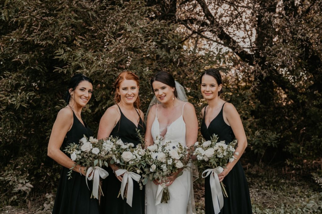 Bride and three bridesmaids in black gowns stand closely together and smile for a formal wedding portrait. Image is cropped knees-up. Captured on the grounds of Cellar 52 in St Jacobs, ON by top Ontario wedding photographer Ashlee Ellison.