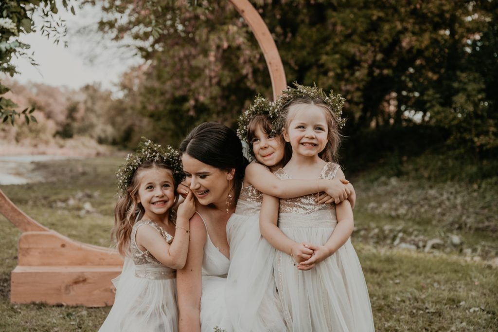 Bride crouches down and is laughing with her three flower girls. Flower girls are in cream and gold dresses with baby's breath floral crowns.