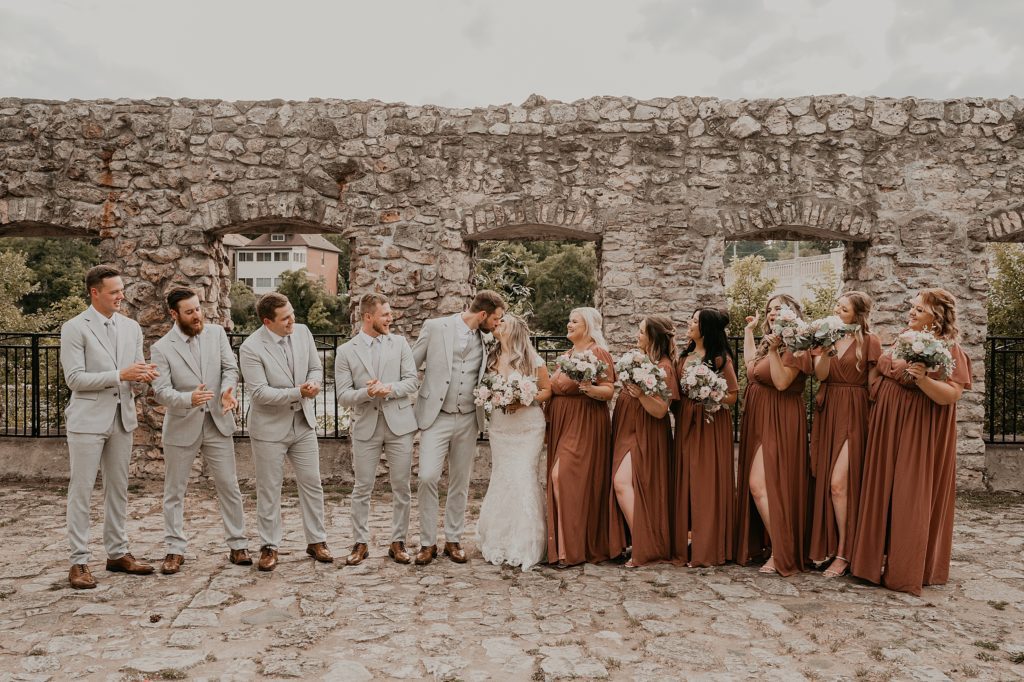 Bride and groom share a kiss at the mill race park ruins as their wedding party stands beside them cheering. Captured by top cambridge ontario wedding photographer ashlee ellison.