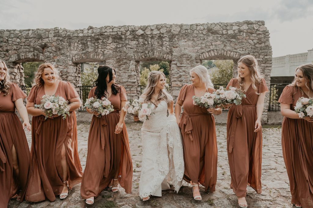 Bride and her bridesmaid are standing in a line and walking amid the ruins at mill race park in cambridge, ontario. the girls are all smiling and laughing together.