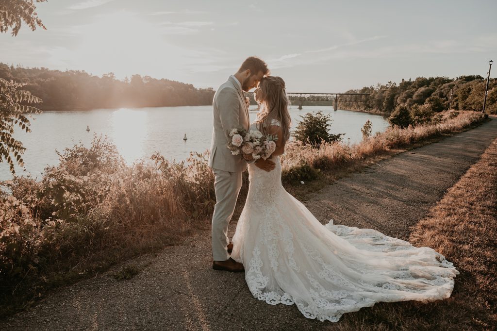 Bride and groom pose for a golden hour portrait at the Cambridge Mill. They are standing in front of the Grand River chest-to-chest with the sun setting and reflecting off the water on the left side of the image. Captured by top Cambridge Ontario wedding photographer Ashlee Ellison.