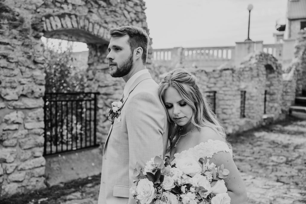 Black and white portrait of bride and groom. Groom's back is to bride and her chest is touching his back. She is resting her head on his back and turned toward the camera. They are standing amid the ruins at mill race park and the Cambridge Mill is seen in the distance. Captured by best wedding photographer in Cambridge Ashlee Ellison.