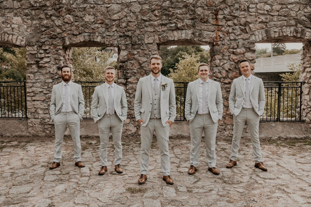 Groom and groomsmen stand with their hands in their pockets at Mill Race Park posing for a formal portrait.