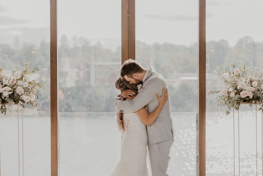 Bride and groom share a hug after they have been announced man and wife at the Cambridge Mill. Large floor-to-ceiling window are behind the couple overlooking the Grand River.
