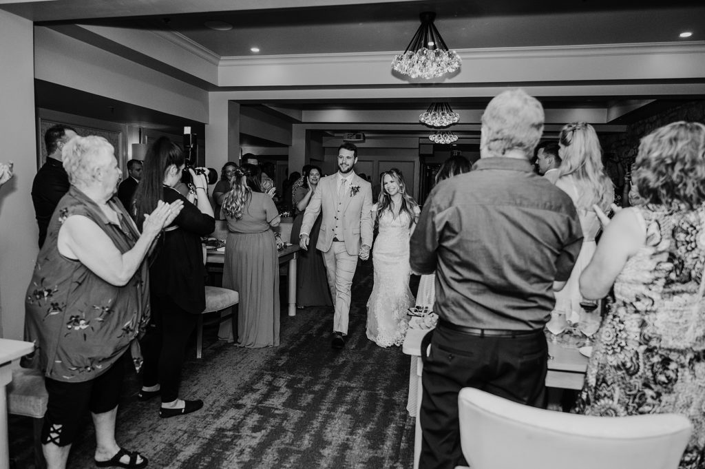 Bride and groom walk hand-in-hand to their Cambridge Mill wedding reception as guests stand and clap.