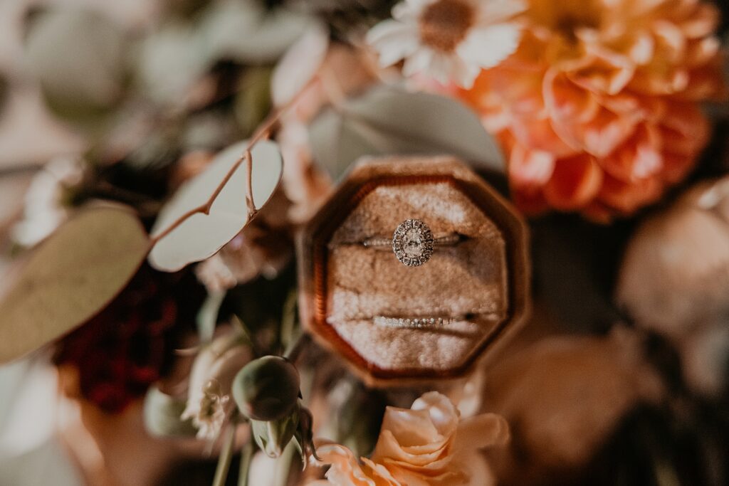 Ring detail shot. Oval engagement ring with a pave halo and eternity-style wedding band are featured in a blush and copper coloured velvet ring box. the box is laid atop of the bride's bouquet of coral, copper, sage, and muted blush hues. Captured by best London, Ontario wedding photographer Ashlee Ellison.