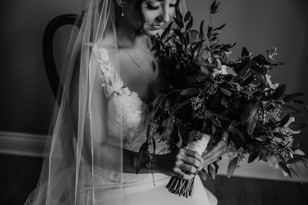 Black and white image of bride posing for portrait after she has just finished getting ready. Bride is sitting on a chair with her veil over her shoulder and holding her bouquet. She is looking down at her flowers and smiling. Captured by top newmarket wedding photographer ashlee ellison.