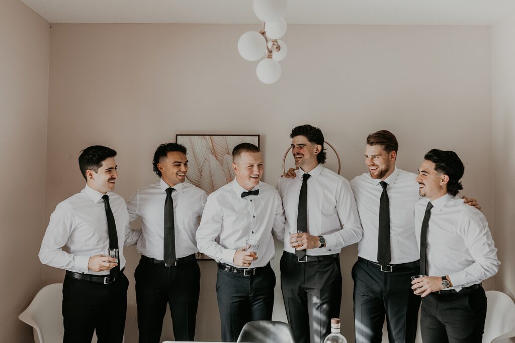 Groom and groomsmen stand in the dining room enjoying a whiskey before his wedding ceremony. Their arms are wrapped around each other and lauging. captured by top ontario wedding photographer ashlee ellison.