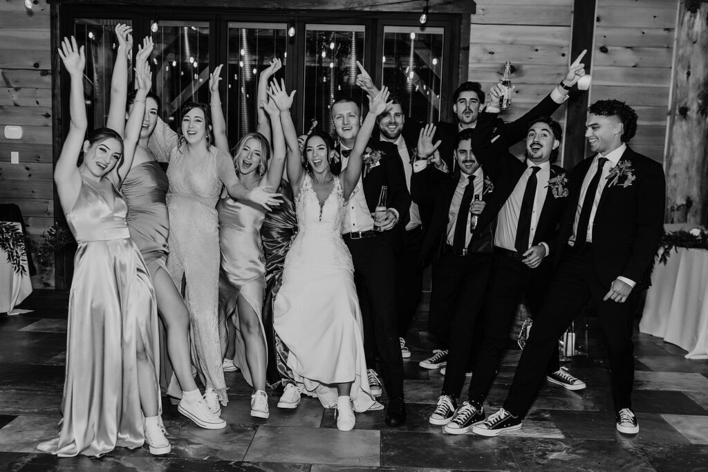 black and white image of bride, groom, and wedding party throw their hands up in celebration as they pose for a casual image at their holland marsh wineries wedding reception. the bride and bridesmaids are wearing white converse, the groom and groomsmen are wearing black converse. captured by best newmarket wedding photographer ashlee ellison.