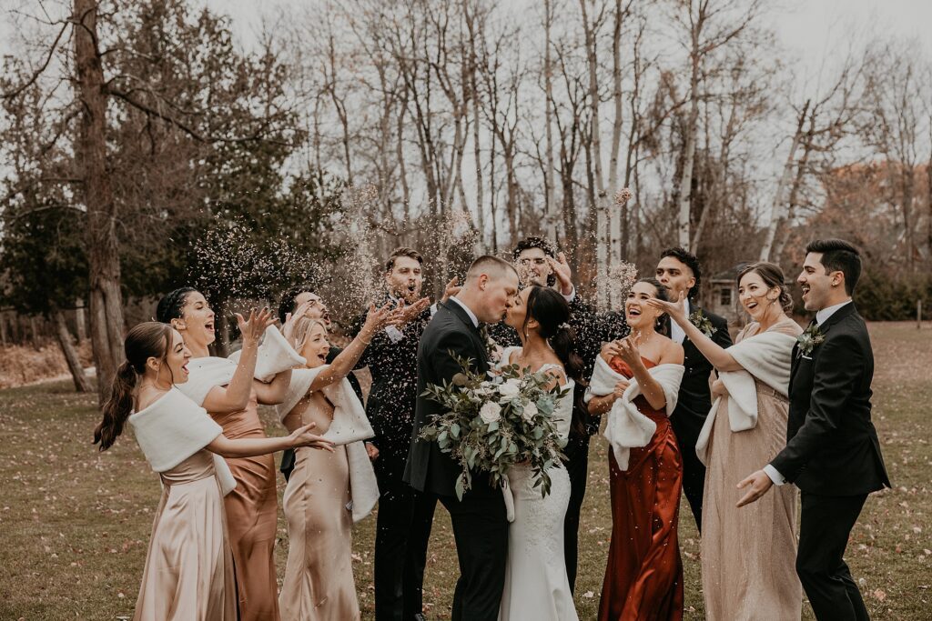 Bride and groom share a kiss as their wedding party throws white confetti on them at their holland marsh wineries wedding. wedding party has a joyful expression on their faces. captured by best newmarket wedding photographer ashlee ellison.